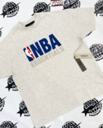 T-shirt grigia NBA Essentials streetwear con logo oversize e sfondo di loghi sportivi urbani.