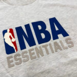 Maglietta grigia NBA Essentials con logo, silhouette di giocatore e stile casual sportivo.