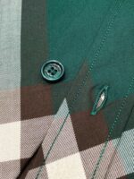 Dettaglio di camicia a quadri con bottone verde lucido e cuciture eseguite a mano.