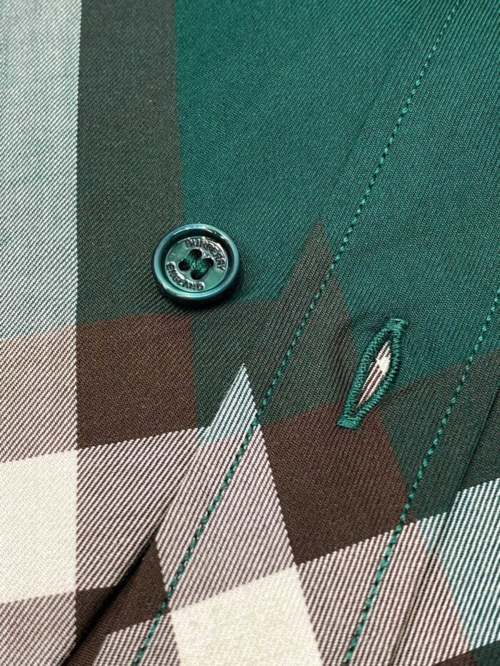 Dettaglio di camicia a quadri con bottone verde lucido e cuciture eseguite a mano.