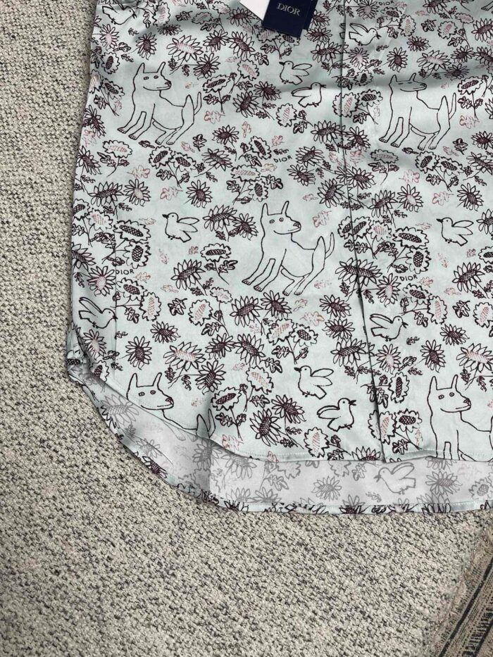 Maglietta azzurra con disegni di cani e fioretti artistici, perfetta per uno stile allegro e casual.