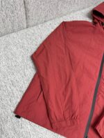 Giacca leggera rossa con cappuccio e zip, su superficie tessile casuale.