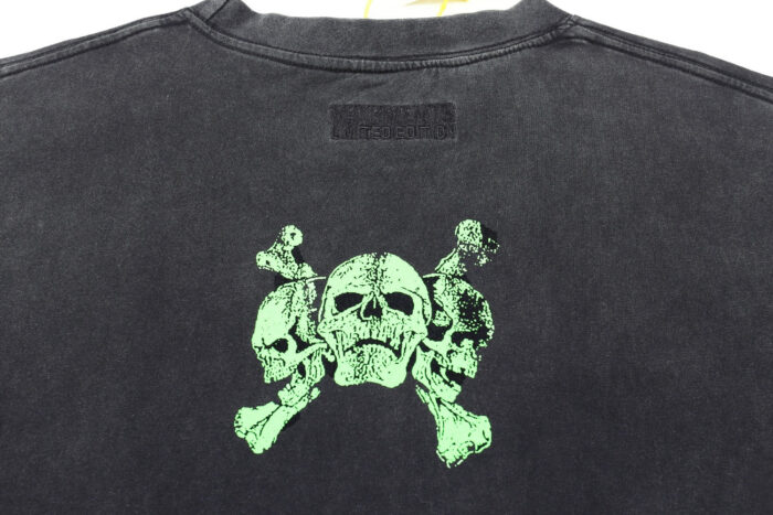 Maglietta nera con grafica di tre teschi verdi e croci dal design distressed, stile punk-rock.