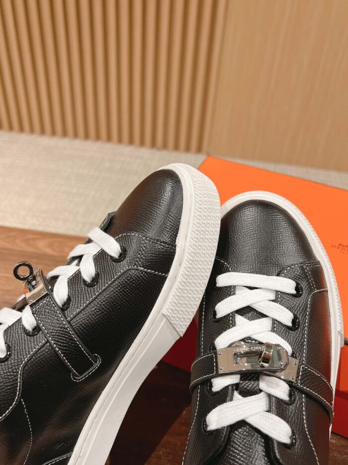 Scarpe sneakers nere in pelle con lacci bianchi e dettaglio metallico, su superficie in legno calda.
