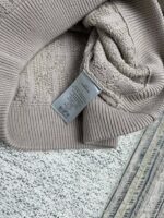 Maglione beige morbido con etichetta, su sfondo a pattern sottile e texture tessile.
