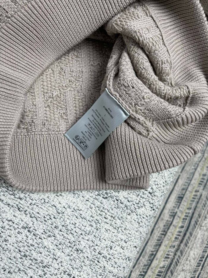 Maglione beige morbido con etichetta, su sfondo a pattern sottile e texture tessile.