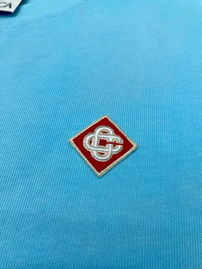 Camicia blu con patch ricamato a diamante rosso e logo geometrico bianco.