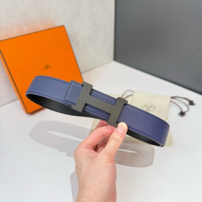 Cintura blu navy con fibbia minimalista di lusso, accompagnata da scatola arancione e custodia elegante.