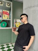 Giovane uomo con occhiali, drink e stile urbano davanti a caffè colorato e muro grigio.