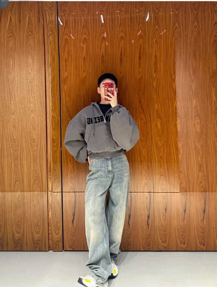 Persona in stile streetwear con hoodie oversized, jeans e sneakers, scatta selfie in ambiente moderno.