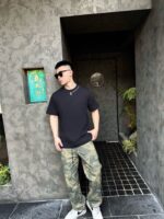 Uomo stile urbano con camo e occhiali, ingresso moderno con dettagli contemporanei.