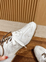Sneaker bianca con lacci marroni su superficie di legno, stile sportivo ed elegante e dettagli curati.