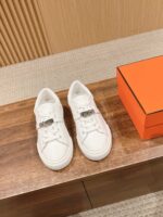 Sneaker bianche di lusso in pelle con dettaglio metallico su superficie in legno, stile elegante e moderno.