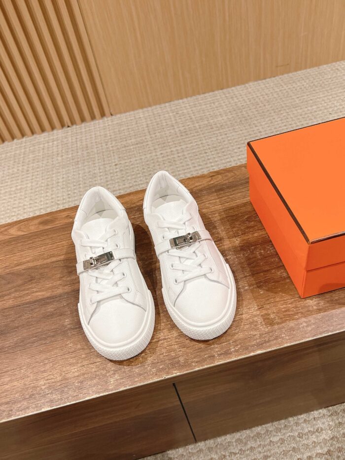 Sneaker bianche di lusso in pelle con dettaglio metallico su superficie in legno, stile elegante e moderno.