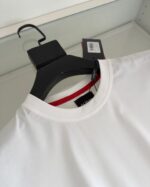 Maglietta bianca minimalista con dettaglio rosso sul colletto e tag, stile casual e versatile.